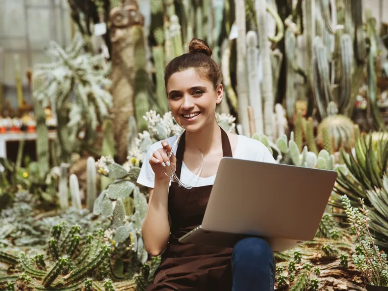 A smiling woman sitting among plants in a greenhouse with a laptop