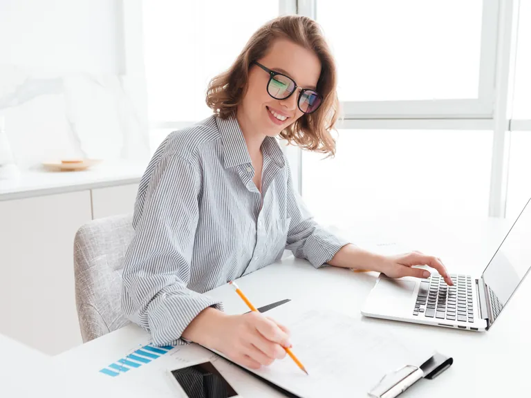 A smiling woman working on a laptop and notes at a bright desk
