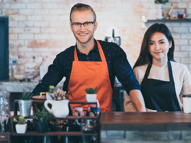 Two smiling small business owners in aprons behind their counter