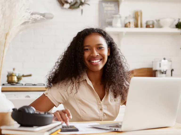 A smiling self-employed woman at a kitchen table with a laptop and calculator