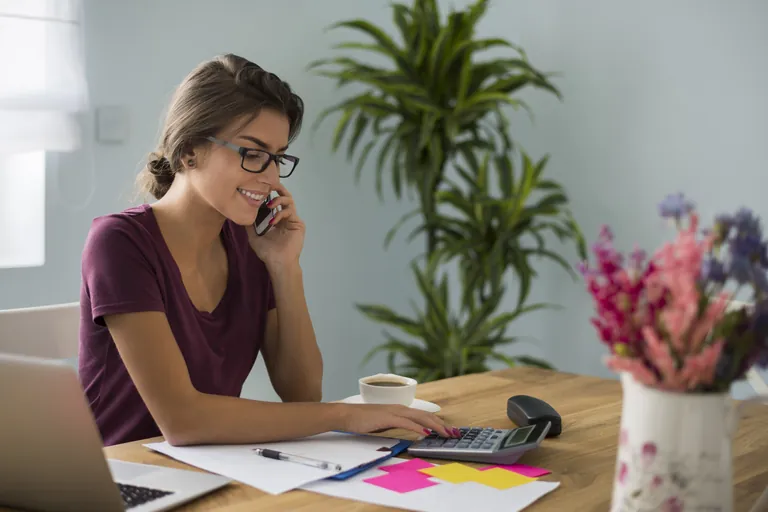 A woman on the phone at her desk with a laptop, calculator and paperwork