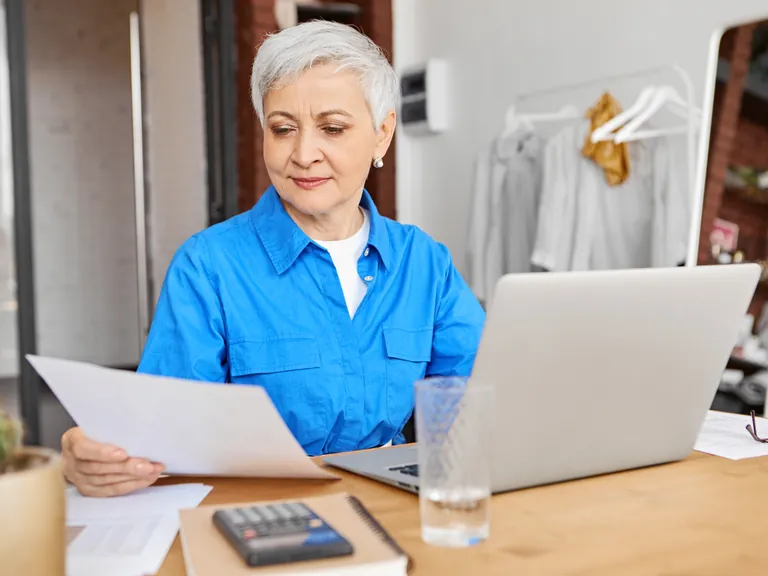 A woman reading a document at a laptop with a calculator on her desk