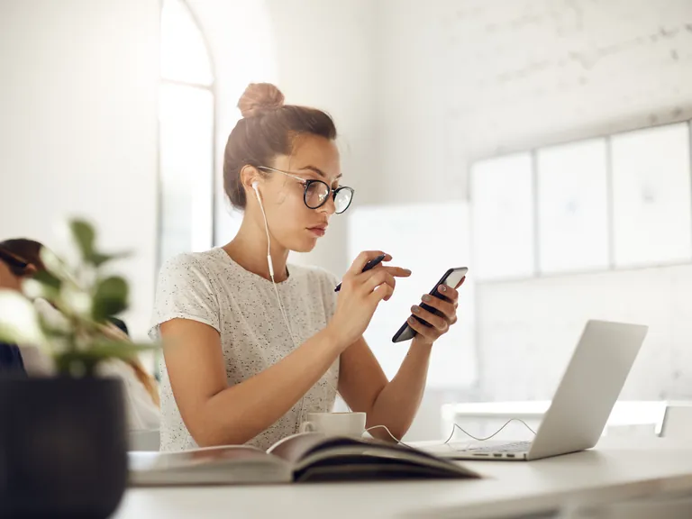 A woman using a smartphone beside her laptop at a bright desk